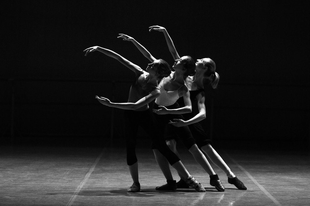 Black and white image of three ballet dancers side by side with their arms extended in a graceful pose